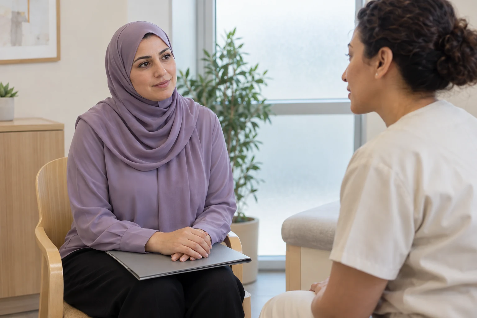 امرأة بملابس وحجاب بنفسجيين تجلس أمام مختصة تحمل زيًا كريميًا، وتضع يديها فوق ملف مغلق في حضنها.