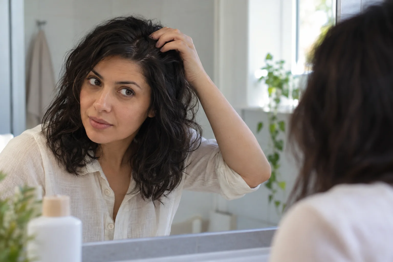 امرأة تنظر في المرآة وتفصل شعرها برفق بأصابعها، بجوار عبوة بلا علامة تجارية.